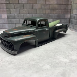 May include: A dark green toy truck model with a weathered, distressed finish showing patches of rust. The vehicle features a front grille, a cab with open windows, and an open truck bed containing a light green cylindrical object.