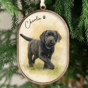 May include: Oval wooden ornament featuring a watercolor painting of a black Labrador puppy with a blue collar. The name "Charlie" is written in script above the puppy, with a paw print symbol. The ornament is hanging from a white ribbon.