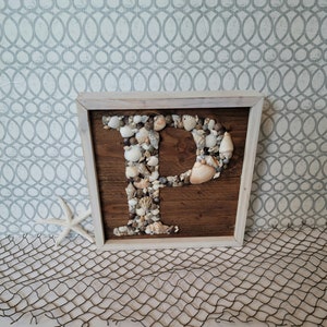 May include: A white wooden frame with a brown wooden background. The letter "P" is made of seashells and is displayed in the center of the frame.