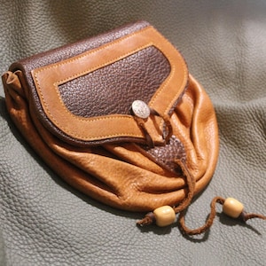 May include: A brown leather pouch with a flap closure and a decorative metal button. The pouch has a drawstring closure and is decorated with wooden beads.