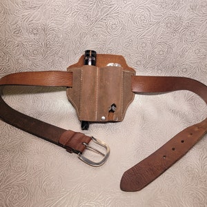 May include: A brown leather belt with a brown leather pouch attached. The pouch has two slots for carrying items. The belt has a silver buckle.