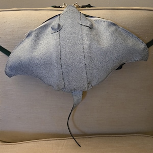 May include: A stingray-shaped bag featuring a textured, speckled grey and white surface. The bag has a wide, flat body with pointed side fins, a central seam, and a long, thin tail extending from the bottom. A silver zipper is visible at the top.