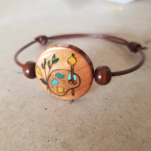 May include: A wooden bracelet with a round wooden charm featuring a painted image of an artist's palette with paintbrushes and a sprig of green leaves.