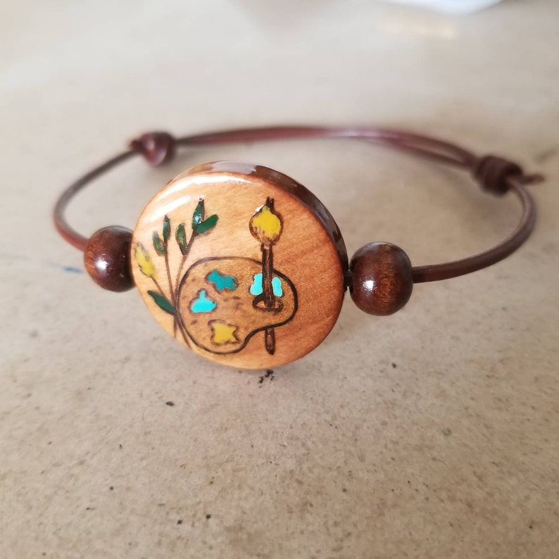 May include: A wooden bracelet with a round wooden charm featuring a painted image of an artist's palette with paintbrushes and a sprig of green leaves.