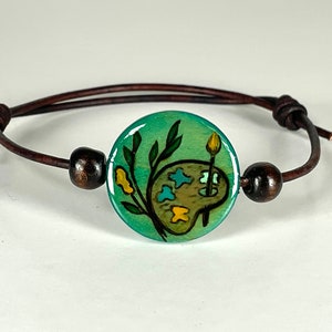 May include: A brown leather bracelet with a round, green, painted ceramic charm featuring a paint palette and a paintbrush. The charm has a green background with yellow and blue accents.