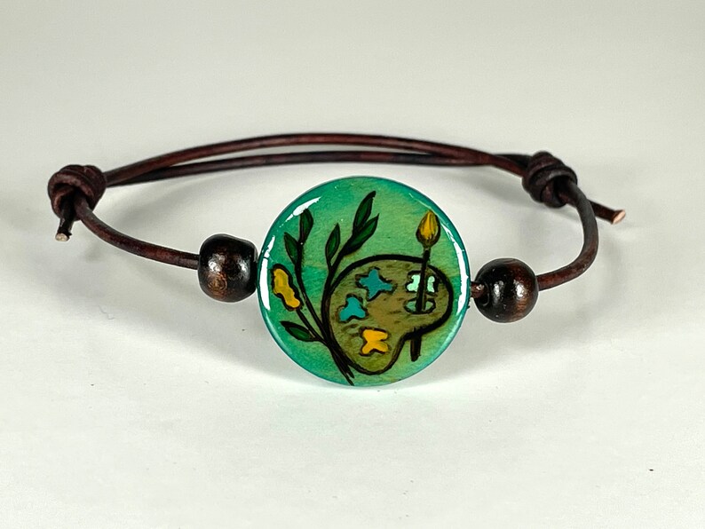 May include: A brown leather bracelet with a round, green, painted ceramic charm featuring a paint palette and a paintbrush. The charm has a green background with yellow and blue accents.