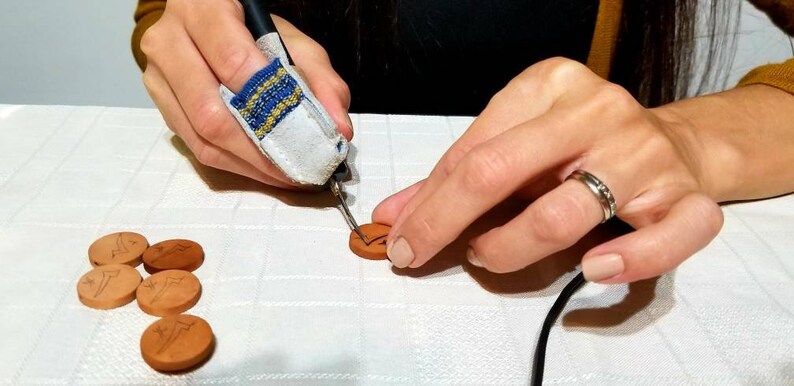May include: A person is using a wood burning tool to create designs on small, round, terracotta tiles. The tiles have a design of a bird on them.