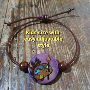 May include: A purple wooden bracelet with a painted palette and paintbrush design. The bracelet is made with brown leather cord and wooden beads. "Kids size with easy adjustable style" is written on the image.