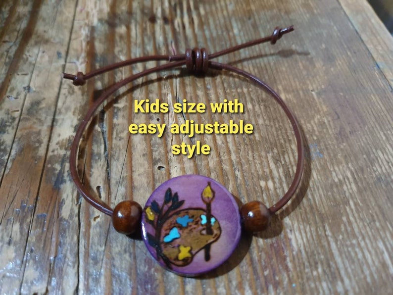 May include: A purple wooden bracelet with a painted palette and paintbrush design. The bracelet is made with brown leather cord and wooden beads. "Kids size with easy adjustable style" is written on the image.