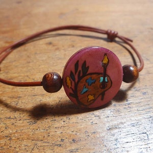 May include: A brown leather bracelet with a wooden bead and a round wooden charm. The charm is painted with a pink background and a stylized image of a paint palette and paintbrush.