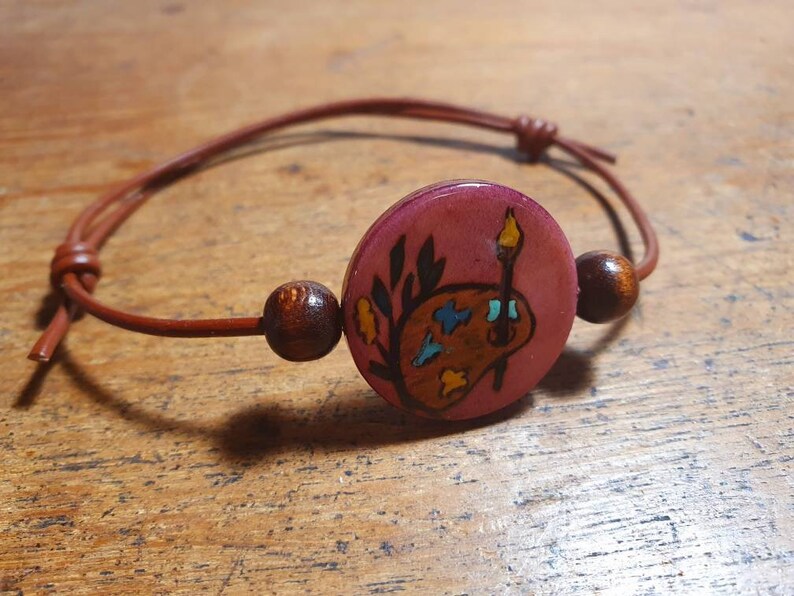 May include: A brown leather bracelet with a wooden bead and a round wooden charm. The charm is painted with a pink background and a stylized image of a paint palette and paintbrush.