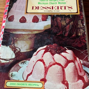 May include: A vintage cookbook titled "Favorite Recipes From Wesleyan Church Women Desserts Including Party Beverages" with a pink dessert topped with red sauce and strawberries on the cover. The book has a red and white checkered binding.