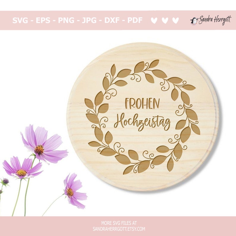 May include: A circular wooden plaque featuring a leaf wreath design and the German text "FROHEN Hochzeitstag" engraved in the centre. The plaque is light brown with a natural wood grain. The image also includes three pink flowers.