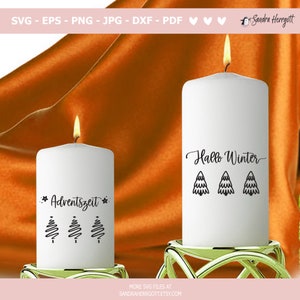 May include: Two white pillar candles with lit flames. One candle has the text "Adventszeit" and three stylized Christmas trees. The other candle has the text "Hallo Winter" and three stylized Christmas trees. The candles are on gold candle holders.
