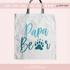 May include: A white tote bag with black handles features the words "Papa Bear" in teal script and a teal bear paw print. The top of the bag has the text "SVG - EPS - PNG - JPG - DXF". The bottom of the bag has the text "MORE SVG FILES AT ETSY.COM/AU/SHOP/SANDRAHERRGOTT".
