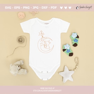 May include: A white baby onesie with a line drawing of a fox holding a star on a crescent moon. The onesie is surrounded by a teddy bear, a star, and a string of wooden beads.