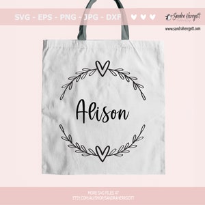 May include: A white tote bag featuring a black design of a wreath with leaves and hearts, and the name "Alison" in the centre.