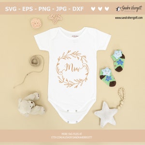 May include: A white baby onesie with a gold floral wreath and the name "Mia" in a cursive font.
