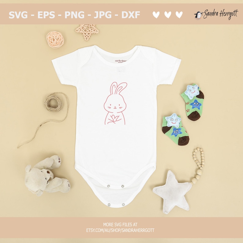 May include: A white baby onesie with a pink outline of a bunny holding a flower. The onesie is surrounded by a variety of baby items, including a star-shaped rattle, a teddy bear, and a pair of socks with stars.