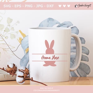 May include: White ceramic mug featuring a pink bunny silhouette and the words "Mama Hase" in a script font. The mug has a curved handle and is likely used for beverages. The design suggests a theme related to Easter or motherhood.