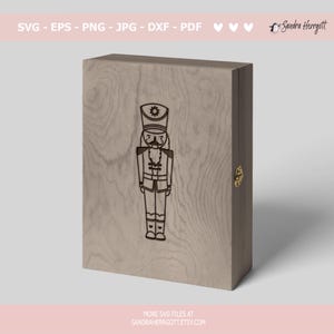 Może przedstawiać: Drewniana skrzynka z grawerowanym wzorem na froncie przedstawiającym łamacza orzech&oacute;w. Skrzynka ma złoty zamek i jest wykonana z jasnobrązowego drewna.