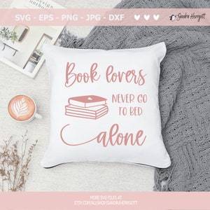 以下が含まれることがあります： ピンクの文字で「Book lovers never go to bed alone」と書かれた白い枕。文字は筆記体。ピンクの3冊の本が積み重ねられており、一番上の本には小さなハートマークが描かれている。枕は灰色の毛布の上に置かれている。