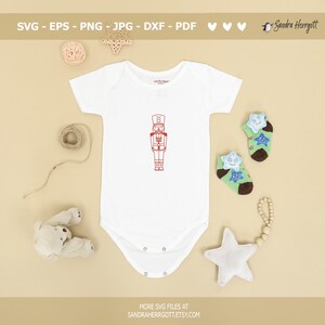 Może przedstawiać: Biały śpioszek dla niemowlęcia z czerwoną ilustracją postaci Dziadka do Orzech&oacute;w z przodu. Śpioszek leży na beżowej powierzchni z brązową kulką wł&oacute;czki, białą gwiazdą i pluszowym misiem.