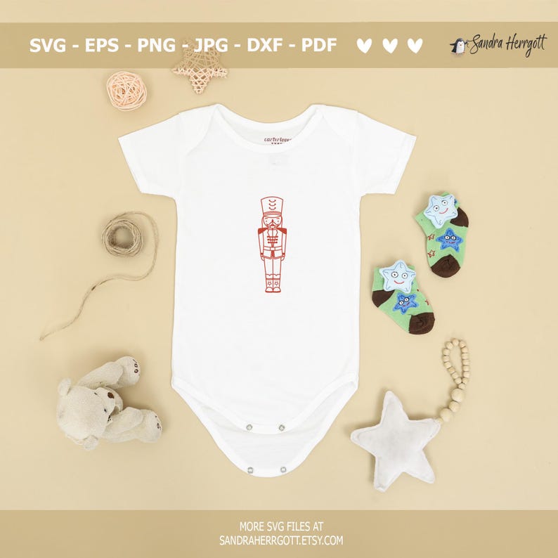 Może przedstawiać: Biały śpioszek dla niemowlęcia z czerwoną ilustracją postaci Dziadka do Orzech&oacute;w z przodu. Śpioszek leży na beżowej powierzchni z brązową kulką wł&oacute;czki, białą gwiazdą i pluszowym misiem.