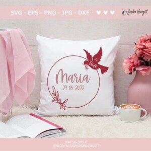May include: A white pillow with a red circle design featuring the name Maria, the date 24-05-2022, and a dove with the word "Taufe" written on it. The pillow is on a light brown surface with a book and a cup of coffee.
