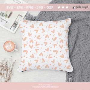 May include: A white pillow with a repeating pattern of peach-colored hearts and dots. The pillow is on a light gray surface with a gray blanket in the background.