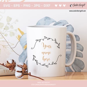 May include: White ceramic mug with a black outline of a floral wreath and the text "Your name here" in gold lettering.