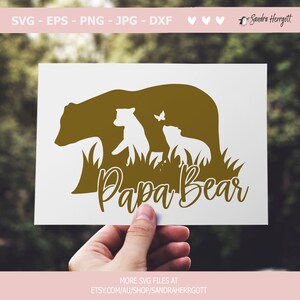 May include: A gold-colored Papa Bear design featuring a bear silhouette with two cubs and a butterfly. The design includes the text "Papa Bear" in a cursive font. The image is held against a blurred green background.