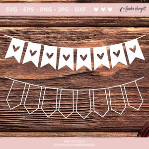 May include: White paper banner with a repeating pattern of flags with a heart cut-out. The banner is hanging from a string of small white triangles. The banner is on a brown wooden background.