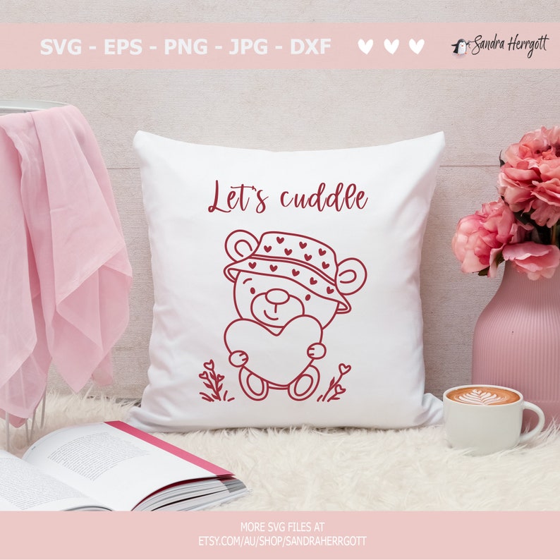 May include: A white pillow with a red outline of a bear wearing a hat with hearts on it. The bear is holding a heart and the text "Let's cuddle" is above the bear.