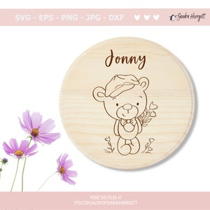 May include: A round wooden sign with the name "Jonny" written in brown cursive lettering. A line drawing of a teddy bear wearing a hat and holding flowers is in the center of the sign.