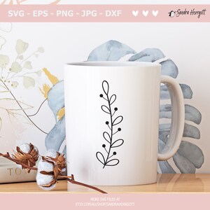 May include: A white ceramic mug with a black line drawing of a simple branch with leaves and berries. The mug is on a wooden surface.