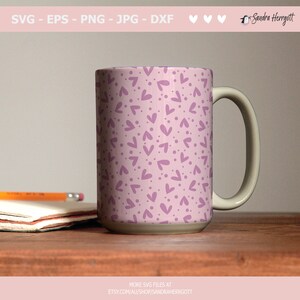 May include: A white ceramic mug with a handle. The mug has a pink background with a pattern of purple hearts and dots.