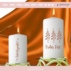 May include: Two white pillar candles with lit flames. One candle has the text "Adventszauber" and star accents. The other candle has three stylized Christmas trees and the text "Frohes Fest" in brown. Both candles sit on gold candle holders.