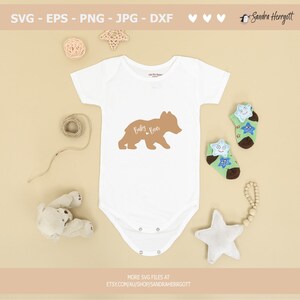 May include: A white baby onesie with a brown bear graphic and the text "Baby Bear." The image includes baby socks, a stuffed bear, a star-shaped decoration, and the text "SVG - EPS - PNG - JPG - DXF." The Etsy shop URL is also visible.