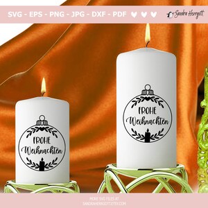 Op de afbeelding: Twee witte stompkaarsen met een zwart kerstornamentontwerp met de tekst "Frohe Weihnachten". De kaarsen zijn aangestoken met een gele vlam en staan op gouden kandelaars. De achtergrond is een gedrapeerde oranje stof.