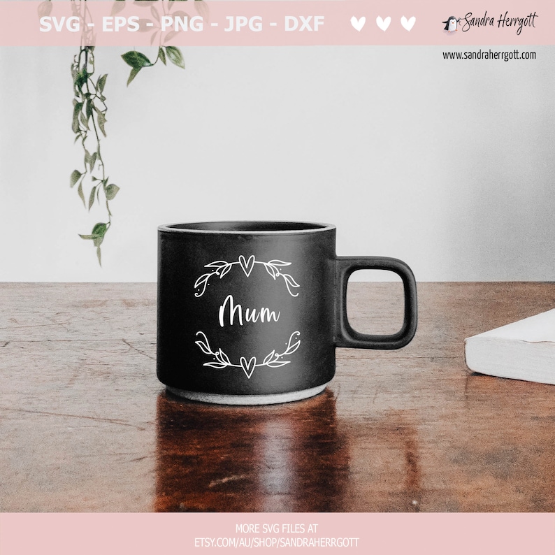May include: A black ceramic mug with a white design of a floral wreath with the word "Mum" in the centre. The mug is sitting on a wooden table.