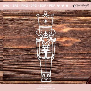 Może przedstawiać: Biały zarys tradycyjnego żołnierza-łamacza orzech&oacute;w z koroną, wąsami i brodą. Łamacz orzech&oacute;w stoi z ramionami opuszczonymi wzdłuż ciała i nosi mundur z guzikami i szarfą.