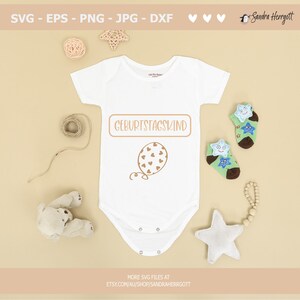 May include: A white baby onesie with the text "GEBURTSTAGSKIND" in a brown rectangle and a brown balloon with hearts on it. The onesie is surrounded by a teddy bear, a star, and a string of wooden beads.