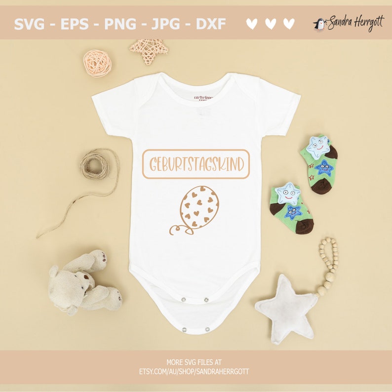 May include: A white baby onesie with the text "GEBURTSTAGSKIND" in a brown rectangle and a brown balloon with hearts on it. The onesie is surrounded by a teddy bear, a star, and a string of wooden beads.