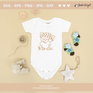 May include: A white baby onesie with a brown line drawing of a teddy bear wearing a hat with hearts on it. The teddy bear is holding a heart in its paws. The onesie is on a beige background with other baby items.