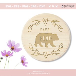 May include: A round wooden sign with the words "PAPA BEAR" and a bear silhouette carved into the wood. The sign is surrounded by a decorative leaf and heart design. The text "SVG - EPS - PNG - JPG - DXF" is at the top.