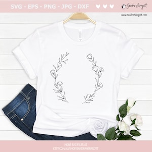 May include: A white t-shirt featuring a black line drawing of a floral wreath. The wreath is composed of leaves and flowers. The t-shirt is displayed on a wooden surface.