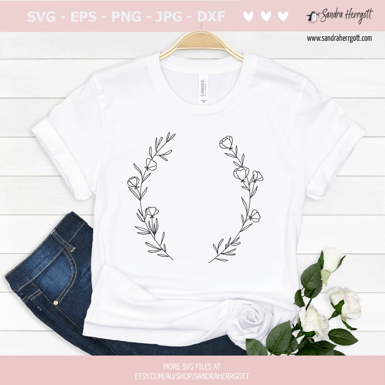 May include: A white t-shirt featuring a black line drawing of a floral wreath. The wreath is composed of leaves and flowers. The t-shirt is displayed on a wooden surface.