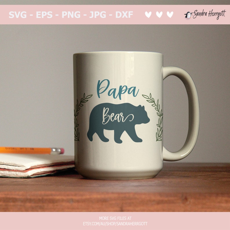 May include: A large, off-white ceramic mug with a teal "Papa Bear" design and a bear silhouette. The mug has a curved handle and is decorated with a leafy wreath. The mug is on a wooden surface.