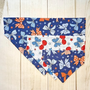 May include: Three triangular pet bandanas featuring a patriotic theme. The fabric displays a repeating pattern of orange, light blue, and white bows interspersed with small stars on dark blue and white backgrounds, with some cherry motifs.